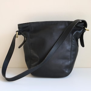 May include: A black leather shoulder bag with a long strap. The bag has a bucket shape and gold-colored hardware. The bag is sitting on a white surface against a neutral background.