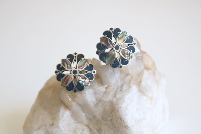 May include: A pair of silver earrings with a blue and iridescent abalone shell design. The earrings are in the shape of a flower with a circular center.