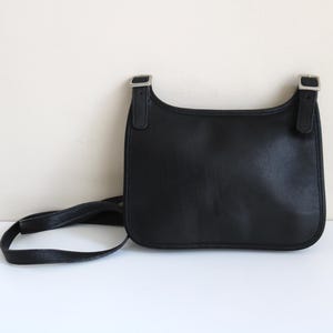May include: Black leather crossbody bag with a rectangular shape and a long, thin strap. The bag features silver-toned buckles on the top corners and a curved top edge. The bag is set against a neutral background.