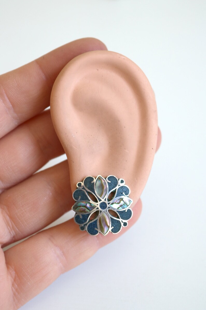 May include: A silver and abalone shell flower-shaped earring with a dark blue background.
