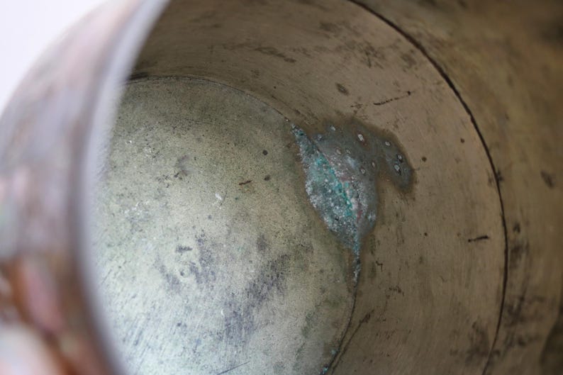 May include: Close-up of the interior of a weathered metal container. The aged surface shows a mix of brown and green hues, with visible signs of wear and oxidation. The container's interior has a textured, aged appearance.