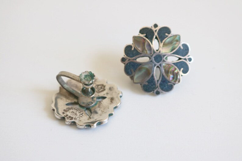 May include: A pair of silver clip-on earrings with a blue and iridescent abalone shell flower design. The earrings are marked "925 Mexico".