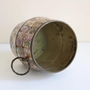 May include: A decorative, barrel-shaped container with a metal rim and handle. The exterior is adorned with iridescent, square-shaped pieces, creating a mosaic-like pattern. The interior has a metallic finish, and the handle is a simple metal ring.