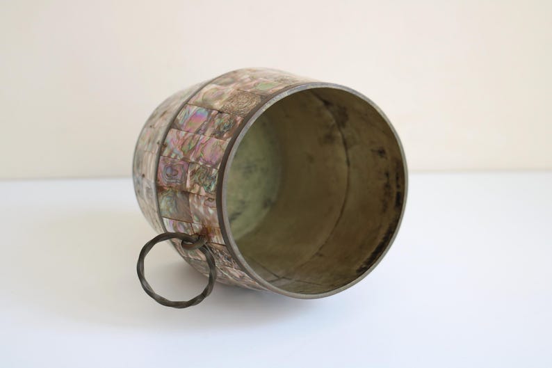 May include: A decorative, barrel-shaped container with a metal rim and handle. The exterior is adorned with iridescent, square-shaped pieces, creating a mosaic-like pattern. The interior has a metallic finish, and the handle is a simple metal ring.