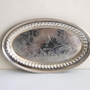 May include: An oval silver-toned serving tray featuring an intricate engraved floral pattern in the center and a decorative rope-style border along the raised rim. The metallic surface has a polished finish with reflective qualities throughout.