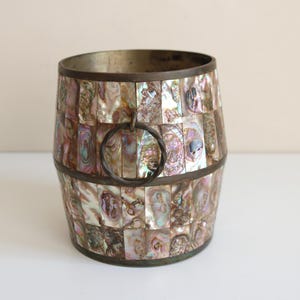 May include: A decorative barrel-shaped container with a mosaic of iridescent abalone shell pieces. The container has a metal rim and a metal handle. The colors include shades of pink, purple, and gold.