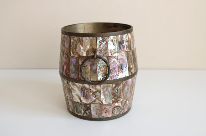 May include: A decorative barrel-shaped container with a mosaic of iridescent abalone shell pieces. The container has a metal rim and a metal handle. The colors include shades of pink, purple, and gold.