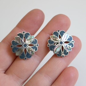May include: A pair of silver earrings with a floral design. The earrings are made of silver and have a blue enamel background. The center of each earring features a flower with iridescent mother-of-pearl petals.