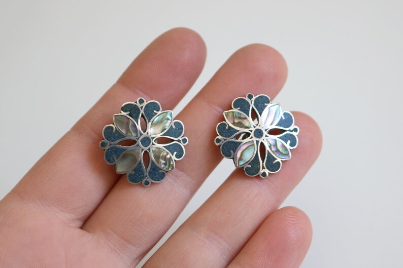 May include: A pair of silver earrings with a floral design. The earrings are made of silver and have a blue enamel background. The center of each earring features a flower with iridescent mother-of-pearl petals.
