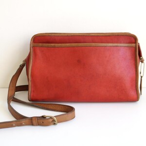 May include: A red leather shoulder bag with a brown leather strap and buckle. The bag has a flap closure and a small pocket on the front.