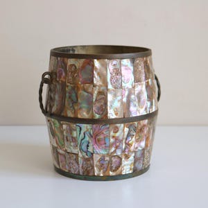 May include: A decorative barrel-shaped container with a mosaic of iridescent, rectangular pieces. The container has a bronze rim and handles. The mosaic pieces display a range of colors, including pink, green, and gold.