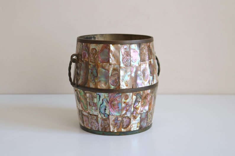 May include: A decorative barrel-shaped container with a mosaic of iridescent, rectangular pieces. The container has a bronze rim and handles. The mosaic pieces display a range of colors, including pink, green, and gold.