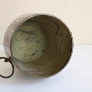 May include: An antique, weathered metal bucket with a dark, circular handle. The interior has a greenish hue, and the exterior shows signs of age and wear. The bucket's shape is cylindrical, and it appears to be made of riveted metal.