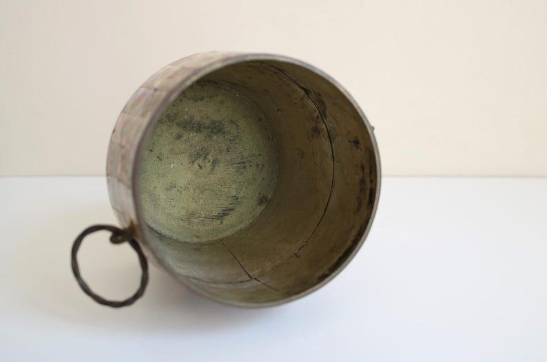 May include: An antique, weathered metal bucket with a dark, circular handle. The interior has a greenish hue, and the exterior shows signs of age and wear. The bucket's shape is cylindrical, and it appears to be made of riveted metal.