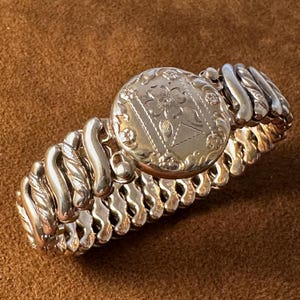 May include: A silver-toned expandable bracelet featuring a central circular medallion with an engraved floral design. The band is composed of repeating, curved, metallic links that create a flexible, stretchable structure for the wrist.