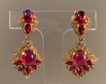 Könnte beinhalten: Ein Paar goldfarbene Ohrhänger mit großen, ovalen und tropfenförmigen roten Edelsteinen. Die Ohrringe sind mit kleineren roten Edelsteinen und klaren Kristallen verziert, die in aufwendigen, filigranartigen Fassungen gefasst sind. Die Ohrringe werden auf einem transparenten Ständer präsentiert.