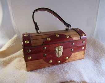 May include: A wooden box purse with a brown leather trim and gold accents. The purse has a top handle and a latch closure.