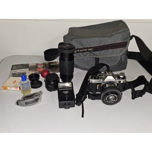 Può includere: Una fotocamera Canon AE-1 argento e nera con obiettivo da 50mm, una borsa fotografica Tamrac, un flash Vivitar, un teleobiettivo, un kit di pulizia per lenti, una piccola pallina rossa, una tracolla e un manuale, disposti su una superficie.