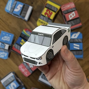 Op de afbeelding: Een wit papieren model van een auto met zwarte accenten en de woorden "Drafty Car" gedrukt op de voorruit. De auto wordt in een hand gehouden.