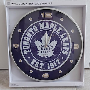 Peut inclure: Une horloge murale ronde avec un cadran bleu marine orné du logo et du texte des Maple Leafs de Toronto. L'horloge a un cadre en métal argenté et affiche les chiffres 12, 3, 6 et 9. Le texte "EST. 1917" est également visible.