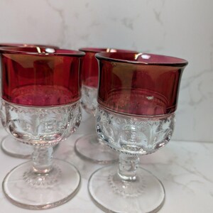 May include: Set of four vintage wine glasses. Each glass features a ruby red bowl and a clear, patterned base. The glasses have a classic design with a rounded bowl and a stemmed foot. The ruby red color adds a touch of elegance.