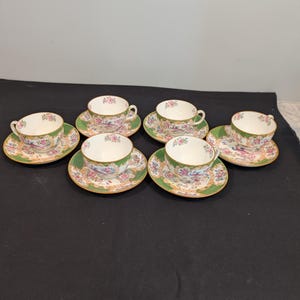 Puede incluir: Seis tazas y platillos a juego. Las tazas y los platillos tienen un fondo blanco con un diseño floral en rosa, amarillo y azul. Los bordes de las tazas y los platillos están adornados con oro, y los platillos tienen un borde verde.