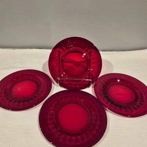 May include: Set of four ruby red glass plates. Each plate has a central, slightly raised circle and a decorative outer rim. The plates are arranged on a white surface, with one plate in a metal stand.