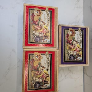 May include: Three wooden boxes with decorative labels. Two boxes have red borders, and one has a purple border. The labels feature a classical artwork with the text "LA PERLA HABANA". The boxes are stacked and arranged against a white background.