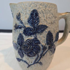 May include: A stoneware pitcher with a textured, light gray surface. The pitcher features a raised design of a rose and leaves in a deep blue color. It has a handle and a spout for pouring.