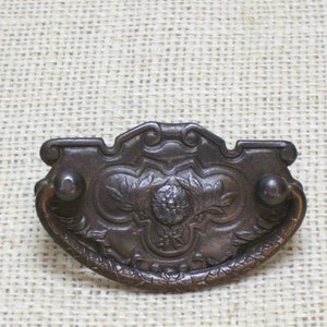 May include: A vintage cast iron drawer pull with an ornate design featuring a floral motif and a central lion's head. The pull has a curved handle and is finished in a dark brown color.