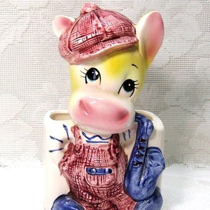 May include: A ceramic planter shaped like a cartoon cow wearing a red and white plaid hat and overalls. The cow is holding a blue saxophone.