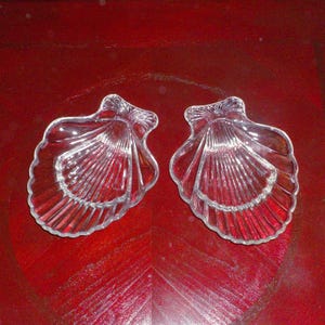 May include: Two clear glass seashell-shaped dishes. Each dish has a scalloped edge and a ribbed interior design. The dishes are displayed on a red surface, creating a contrast with the transparent glass.