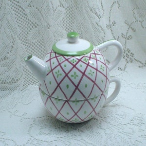 May include: A white ceramic teapot with a green lid and a pink and green geometric pattern. The teapot has a handle and a spout.