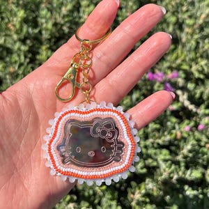 May include: A keychain featuring a reflective, mirror-like Hello Kitty character face cutout. The character is framed by a border of small, vibrant orange beads, which is further surrounded by a scalloped edge of tiny white beads. It has a gold-tone metal ring.