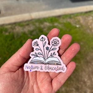 May include: A pink sticker with a floral design and the words "Native & Educated." The sticker features an open book with flowers and leaves sprouting from it. The flowers are in black outline with blue and white accents.
