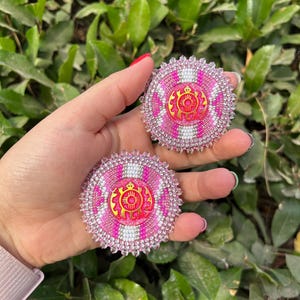 May include: Two round, beaded accessories with a pink, white, and gold design. The center of each accessory features a gold emblem. The edges are lined with clear beads. The accessories are displayed against a background of green foliage.