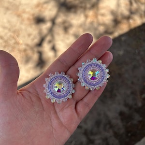 May include: A pair of circular beaded earrings featuring a central iridescent crystal surrounded by concentric rings of lavender and white seed beads, with a scalloped outer edge of clear, light-reflecting beads arranged in a sunburst pattern.