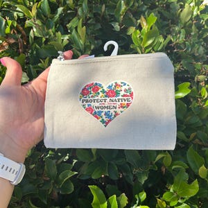 May include: A beige canvas zippered pouch featuring a heart-shaped patch with a colorful floral pattern and the text PROTECT NATIVE WOMEN printed in black letters across the center. The pouch includes a white plastic hanging hook at the top.