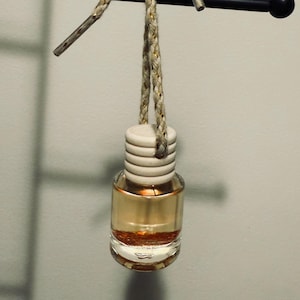 May include: A small, clear glass bottle filled with a golden liquid and suspended by a braided rope with gold accents. The bottle has a wooden cap and is tied with a decorative bow. The product is an air freshener.