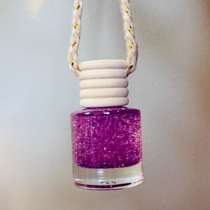 May include: A small, clear glass bottle filled with sparkling purple liquid, topped with a light wood cap and suspended by a cream-colored rope with gold accents. The bottle is likely an air freshener or perfume.
