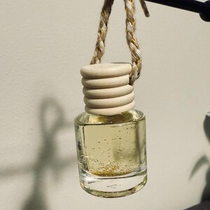 May include: A small, clear glass bottle filled with a golden liquid and gold glitter. The bottle has a wooden cap and is suspended by a braided cord with gold accents. The item is likely an air freshener or fragrance diffuser.