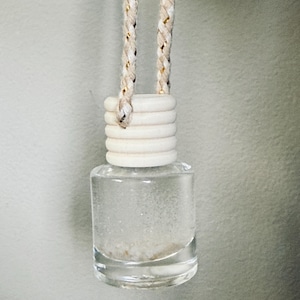 May include: A clear glass bottle with a wooden cap and a braided rope for hanging. The bottle contains a clear liquid with small, white particles. The rope is beige with gold accents. The product is designed for aromatherapy.