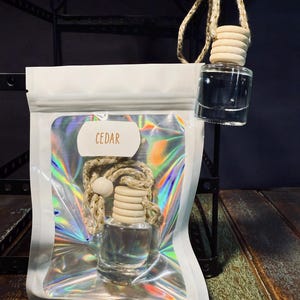 May include: A clear glass bottle with a wooden cap and a string hanging from it. The bottle is labeled "CEDAR" and is packaged in a white plastic bag with a holographic iridescent design.