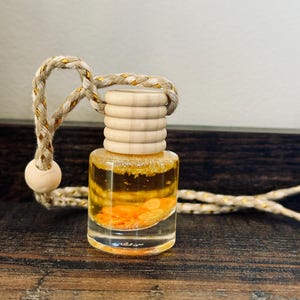 May include: A small, clear glass bottle filled with a yellow liquid and orange particles. The bottle has a wooden cap and a braided rope with gold accents for hanging. The bottle sits on a dark wooden surface.