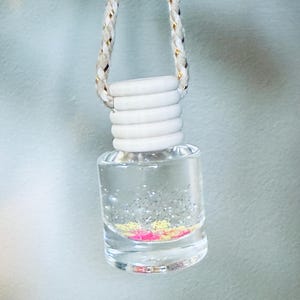May include: A clear glass air freshener bottle with a white cap and a decorative rope. The bottle contains glitter in pink, yellow, and silver. The rope is white with gold accents.