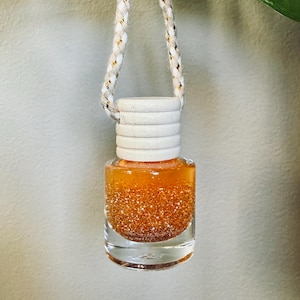 May include: A small glass bottle with a white wooden cap and a gold glitter liquid inside. The bottle is hanging from a white and gold rope.