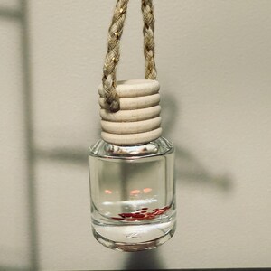 May include: Clear glass car air freshener bottle with a wooden cap and braided rope hanger. The bottle contains a clear liquid with red glitter. The air freshener is hanging against a neutral background.