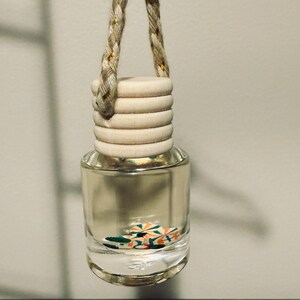 May include: Clear glass car air freshener with a wooden cap and a braided rope for hanging. The air freshener contains colorful decorative pieces. The rope is beige with gold accents.