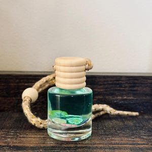 May include: A small, clear glass bottle filled with blue liquid and decorative accents. The bottle has a light wood cap and is attached to a braided rope with a wooden bead. The bottle is sitting on a dark wood surface.