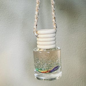 May include: Clear glass car air freshener with a white wooden cap and a silver glitter interior. The air freshener has a rainbow-colored accent at the bottom and is suspended by a beige and gold rope.